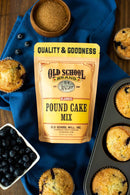 A bag of Old School Brand Pound Cake Mix sits on a blue cloth, surrounded by brown sugar, fresh blueberries, and a muffin pan filled with blueberry muffins.