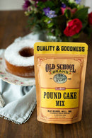 A bag of Old School Brand Pound Cake Mix sits on a wooden table beside a moist, powdered sugar-dusted cake on a plate, with flowers in the background.