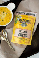 A bag of Old School Brand Stone Ground Yellow Grits sits on a beige cloth beside a bowl of Southern-style grits topped with herbs and cheese, with two silver spoons resting on a dark tray.