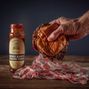 A hand squeezes a messy hamburger with sauce dripping onto red and white checkered paper, next to a jar of Bourbon Barrel Foods Pendennis Club The Original Henry Bain’s Sauce on a wooden table.