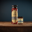 Two jars of Bourbon Barrel Foods Pendennis Club The Original Henry Bain’s Sauce sit on a wooden surface against a dark blue background, each with a cream label featuring blue and gold accents.
