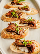 Toasted bread slices topped with creamy cheese, cooked steak, and Bourbon Barrel Foods Pendennis Club The Original Henry Bain’s Sauce, garnished with green herbs and served on a white oval platter.