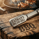 Personalized BBQ Tongs with Name from DDR BBQ Supply rest on a rustic wooden surface next to engraved grilling tools, with a grill and sizzling meat blurred in the background.