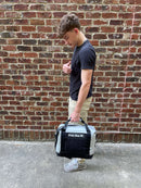 A young man in a black t-shirt, khaki pants, and gray sneakers stands sideways against a brick wall, holding a Polar Bear Coolers 12 Pack Original Nylon Soft Cooler in one hand and giving a thumbs up with his other hand.