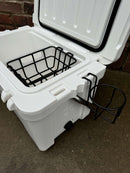 The Polar Bear Coolers' Polar Bear 20 Hard Cooler, in white with an open lid, shows a black wire basket inside and a black wire cup holder attached to the side, placed on concrete with a brick wall in the background.