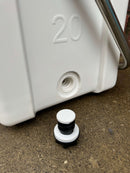 A close-up of a Polar Bear Coolers Polar Bear 20 Hard Cooler, featuring a white body with a threaded hole near the bottom and a black-and-white stopper placed on the ground in front.