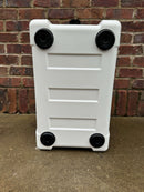 A Polar Bear Coolers Polar Bear 20 Hard Cooler, resembling a white suitcase with four black wheels, rests on its back on concrete. A red brick wall forms the backdrop, highlighting its sleek, durable design and reliable ice retention.