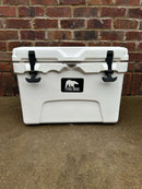 A white Polar Bear 20 Hard Cooler by Polar Bear Coolers with black latches sits on concrete before a red brick wall. The front shows the brand’s bear logo, highlighting its superior ice retention for all your outdoor adventures.