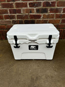 A Polar Bear Coolers 20 Hard Cooler with black latches is on a concrete surface by a red brick wall. The polar bear logo and "COLD TOUGH COOLERS" highlight its excellent ice retention.