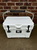 The Polar Bear Coolers Polar Bear 20 Hard Cooler, featuring a white exterior, metal handle with black grip, black latches, and front logo, sits on concrete before a red brick wall. It's known for excellent ice retention.