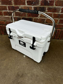 A white Polar Bear 20 Hard Cooler by Polar Bear Coolers with a metal handle and black rubber grips sits on concrete before a red brick wall. It has two black latches, a black logo, and is renowned for excellent ice retention.