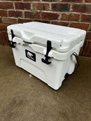 A white Polar Bear 20 Hard Cooler by Polar Bear Coolers, featuring black latches and a handle, sits on concrete before a red brick wall. The front logo showcases this cooler’s superior ice retention performance.