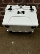 The Polar Bear Coolers Polar Bear 20 Hard Cooler, featuring black latches, wheels, and a logo, stands on a concrete surface before a red brick wall—designed for outstanding ice retention on all your adventures.