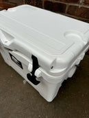 The Polar Bear Coolers Polar Bear 20 Hard Cooler, a white hard-shell cooler with black latches, sits on concrete by a red brick wall. It features cup holders, a textured top, and is designed for maximum ice retention.