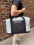 A person in a black shirt and light pants carries a Polar Bear Coolers 24 Pack Original Nylon Soft Side Cooler with a shoulder strap, standing against a brick wall.