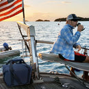 A person in a blue plaid shirt and cap sits on a boat at sunset, drinking from a cup. Behind them waves an American flag, calm water and islands in view, with a Polar Bear 24 Pack Original Nylon Soft Side Cooler by Polar Bear Coolers resting nearby.