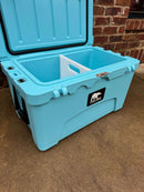 A spacious light blue Polar Bear Coolers 45 Hard Cooler with an open lid sits on concrete by a brick wall. Featuring a white divider, long-lasting ice retention, and the signature black bear logo on the front.
