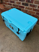 A spacious turquoise Polar Bear Coolers 45 Hard Cooler with black latches and molded cup holders sits on wet concrete before a brick wall, showcasing its rugged design and long-lasting ice retention for outdoor adventures.