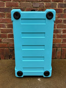 A large, light blue hard-shell suitcase with four black wheels stands upright on wet concrete before a brick wall, echoing the tough resilience of the Polar Bear Coolers' Polar Bear 45 Hard Cooler.