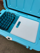 An open Polar Bear 45 Hard Cooler by Polar Bear Coolers sits on concrete with a black wire basket on the left and a handled white cutting board on the right—ideal for rugged outdoor use and long-lasting ice retention.