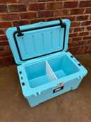 A large light blue Polar Bear 45 Hard Cooler by Polar Bear Coolers sits on concrete with its lid open, revealing a removable divider that splits the rugged cooler into two compartments against a brick wall.