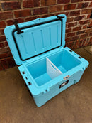 A Polar Bear Coolers Polar Bear 45 Hard Cooler in light blue, featuring extended ice retention and a white divider inside, sits open on concrete against a red brick wall.