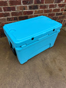 A Polar Bear 45 Hard Cooler by Polar Bear Coolers, featuring a closed lid and bright blue color, rests on a concrete surface before a red brick wall, designed for extended ice retention on any adventure.