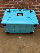 A Polar Bear Coolers 45 Hard Cooler with durable black latches and wheels sits on concrete before a red brick wall, featuring the brand’s black and white logo on top.