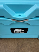 Close-up of a light blue Polar Bear Coolers Polar Bear 45 Hard Cooler with a metal latch and a black-and-white polar bear logo sticker, set on a concrete surface. This rugged outdoor cooler is built for durability.