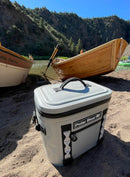 A Polar Bear Coolers Topper 30 Floating Soft Cooler in gray rests on sandy ground by a river, with wooden boats along the shore and green, forested hills under a clear blue sky.