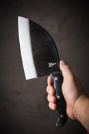 A hand holding the Folded Steel Primrose Serbian Chef Knife with a 1095 high-carbon steel blade and black handle, set against a dark, blurred background—perfect for outdoor cooking.