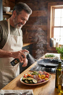 A man in an apron smiles as he uses the Proud Grill Spritz It BBQ Marinade Spray Italian Herb on grilled veggies and meat in a rustic kitchen with wooden walls and sunlight streaming through the window.