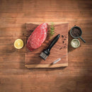 Raw steak on a wooden board with the Amazon Round 24-Blade Meat Tenderizer, half a lemon, rosemary, a brush, black peppercorns, and coarse salt on a wooden table.
