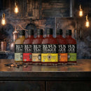 Six bottles of Rufus Teague BBQ Sauce Variety Pack from Rufus Teague are lined up on a rustic kitchen counter with hanging lights and a smoky, industrial backdrop—a perfect BBQ sauce set for any grill master.
