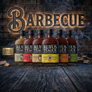 Five bottles from the Rufus Teague BBQ Sauce Variety Pack by Rufus Teague are displayed on a rustic wooden table with a glowing BARBECUE sign and brick wall behind them, next to a small BBQ Foundry box.