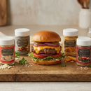 A juicy cheeseburger topped with lettuce, tomato, and red onion is served on a wooden board beside four jars of SALTOPIA Infused Sea Salts from the Saltopia Burger Bundle – the best seasonings for burgers. Shredded cheese and herbs complete the scene.