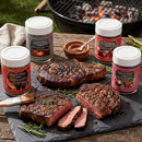 Grilled steaks on a slate board with rosemary, jars of SALTOPIA Infused Sea Salts’ Steak Bundle - Searingly Great Flavors, seasoning bowl, brush, and a backyard grill for true steakhouse-style flavor.