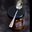 A close-up of a spoon with a small cartoon eye on its handle rests atop a Spice Witch Black Garlic Chili Crisp jar, its black lid visible. The setup hints at tasting Spice Witch’s rich, flavorful condiment against a dark background.