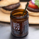 An open jar of Spice Witch Black Garlic Chili Crisp by Spice Witch, an umami condiment, sits with a spoon inside in front of two lettuce and tomato-topped burgers on a wooden board.