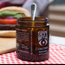 An open jar of Spice Witch Black Garlic Chili Crisp by Spice Witch, a vegan and gluten-free condiment made with avocado oil, sits on a red checkered cloth. The label is visible, with a blurred burger in the background.