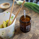 A container of sliced cucumbers with chopsticks and a jar of Spice Witch Black Garlic Chili Crisp, a rich umami condiment by Spice Witch made with avocado oil, sit on a wooden surface with green onions in the background.