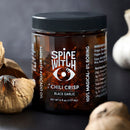 A jar of Spice Witch Black Garlic Chili Crisp by Spice Witch, an umami condiment made with avocado oil, sits on a dark surface among garlic bulbs. The black-lidded jar shows the chili crisp and features white text and a stylized eye logo.