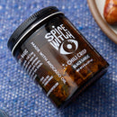 A Spice Witch Black Garlic Chili Crisp jar, an umami condiment from Spice Witch made with avocado oil, rests on its side on a blue textured surface next to a plate of food.