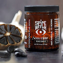 A jar of Spice Witch Black Garlic Chili Crisp stands beside split black garlic, its dark cloves visible. The Spice Witch label features avocado oil and calls this umami-packed condiment “100% magical – 0% boring.”.