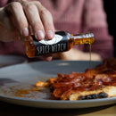 A hand pours Spice Witch Chili Oil from a small bottle onto slices of pepperoni pizza on a plate.