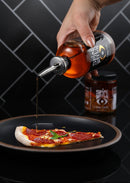 A hand pours Spice Witch Chili Oil from a bottle onto pepperoni pizza on a black plate, enhancing the flavor. A jar of Spice Witch spice mix sits in the background against a tiled wall.