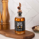 A bottle of Spice Witch Chili Oil by Spice Witch, featuring a large eye graphic on the label and a convenient pour spout, sits on a wooden board with kitchen items and a marble backsplash in the background.