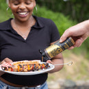 A smiling person holds a plate of grilled skewers and corn while another hand pours Spice Witch Hot Honey from a Spice Witch squeeze bottle onto the food outdoors.