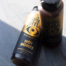 Two Spice Witch Hot Honey Squeeze Bottles rest on a gray surface—one sharp, one blurred. The black and gold label with an eye and "100% Magical, 0% Boring" slogan makes this product perfect for fans of spicy honey. Brand: Spice Witch.
