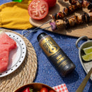 A bottle of Spice Witch Hot Honey Squeeze Bottle sits on a blue picnic blanket surrounded by watermelon slices, lime wedges, tomato, grilled meat skewers, and vegetables.